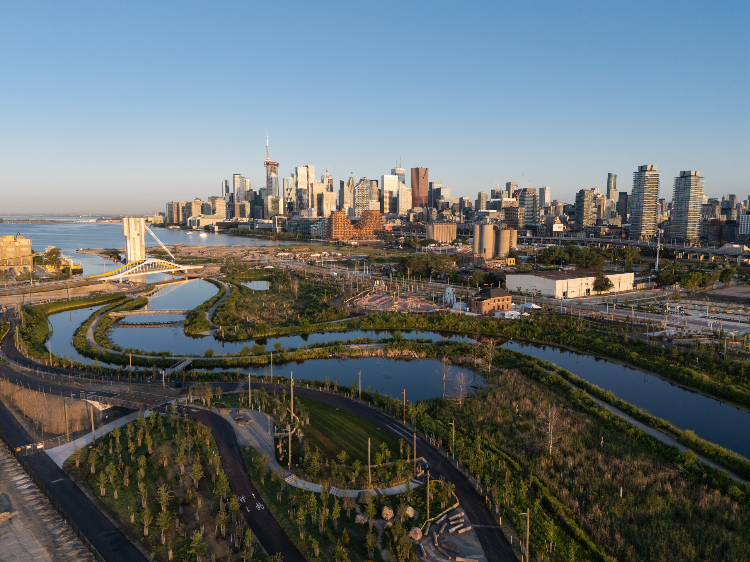 Aerial view of Toronto’s skyline, including the CN Tower, beyond a landscaped waterfront park with winding ponds, walking paths, roads, and a modern pedestrian bridge.