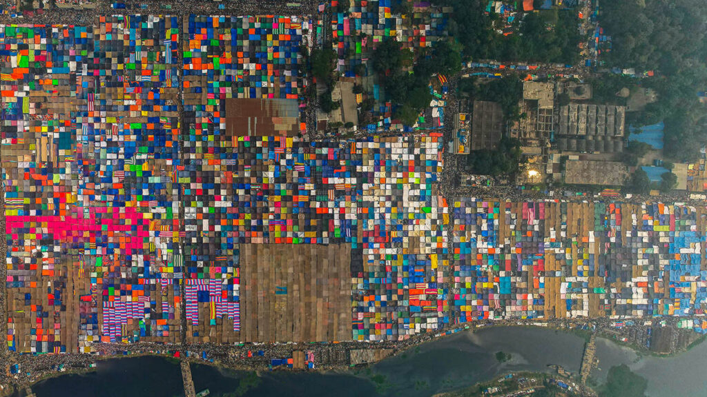 A top-down view of a colorful grid of square tents betweem a city and a river.