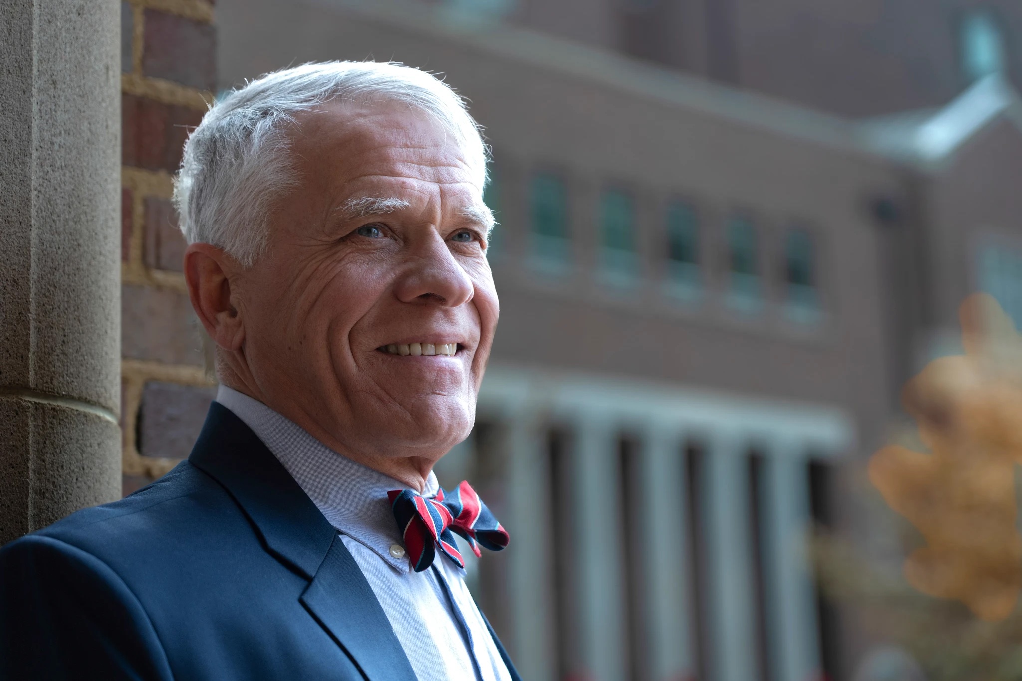 Glenn Mueller, a smiling older man with short white hair wears a dark suit and a red-and-blue striped bow tie, posed beside a brick wall with a softly blurred building in the background.