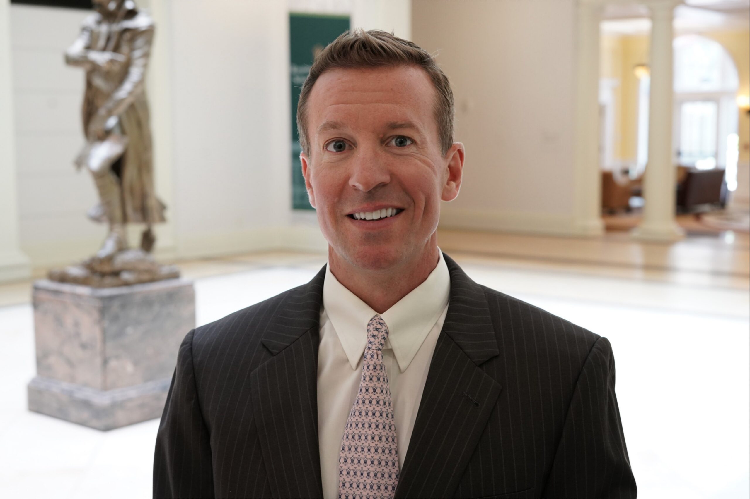 Michael Seiler, a smiling man in a dark pinstripe suit, white shirt, and patterned tie poses in a bright formal interior, with a sculpture and columns softly blurred in the background.