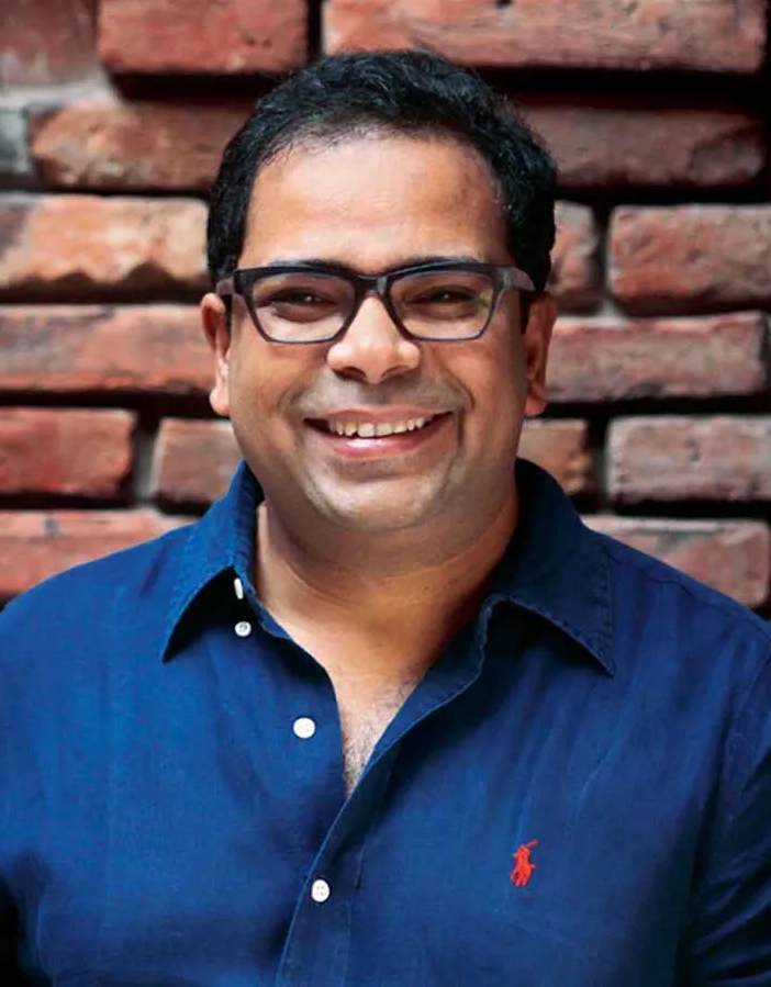 Kashef Chowdhury, wearing dark-framed glasses and a navy blue button-down shirt poses in front of a red brick wall.