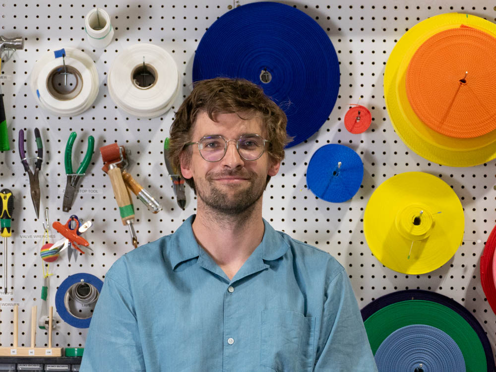 August Lehrecke, Pneuhaus co-founder wearing glasses and a light blue shirt poses in front of a workshop pegboard covered with hand tools and brightly colored rolls of material in blue, yellow, orange, green, and red.