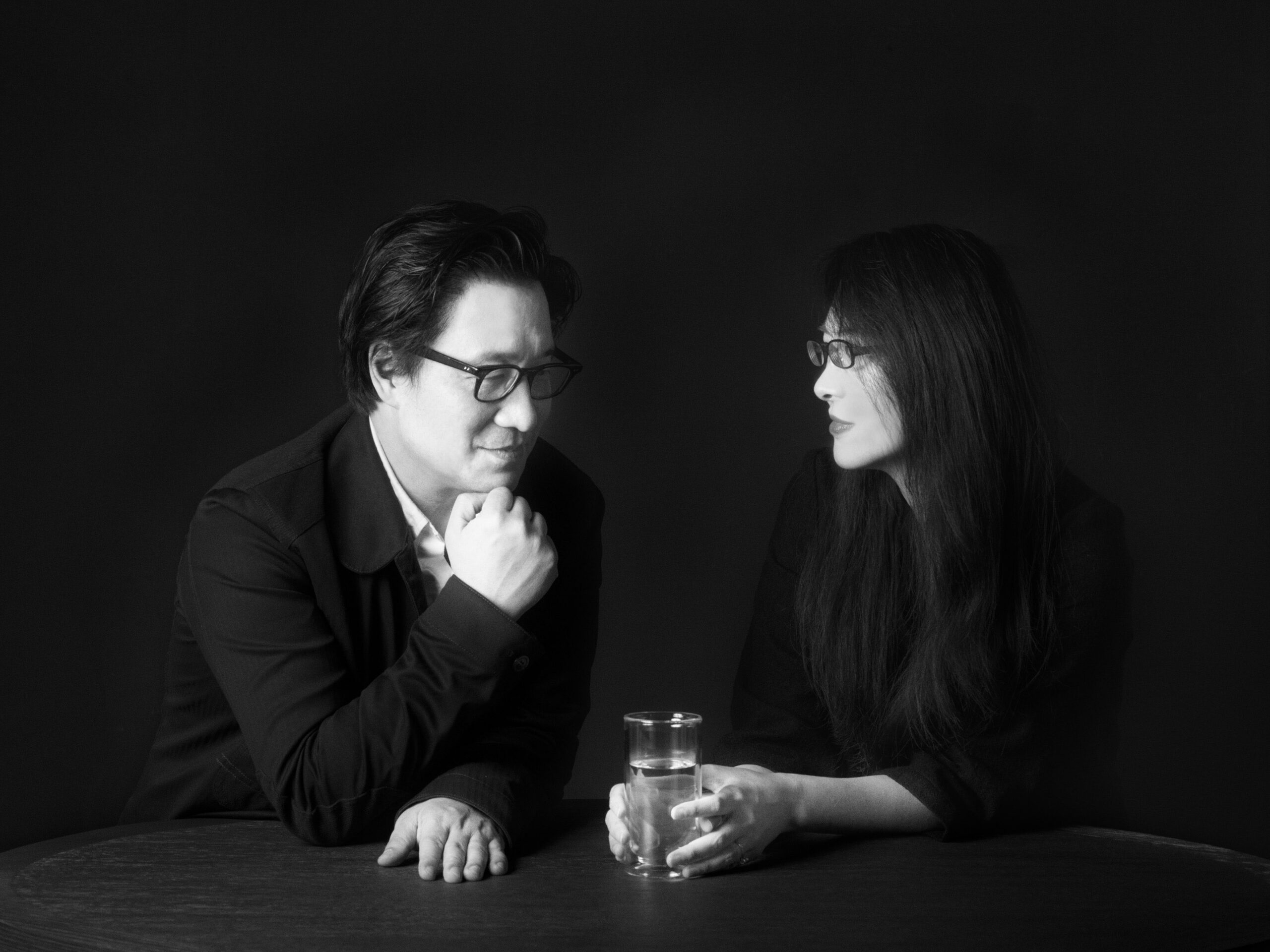 A black-and-white portrait of Lyndon Neri and Rossana Hu wearing glasses seated at a table against a dark background. One rests their chin on their hand while looking toward the other, who holds a glass of water as they face each other.
