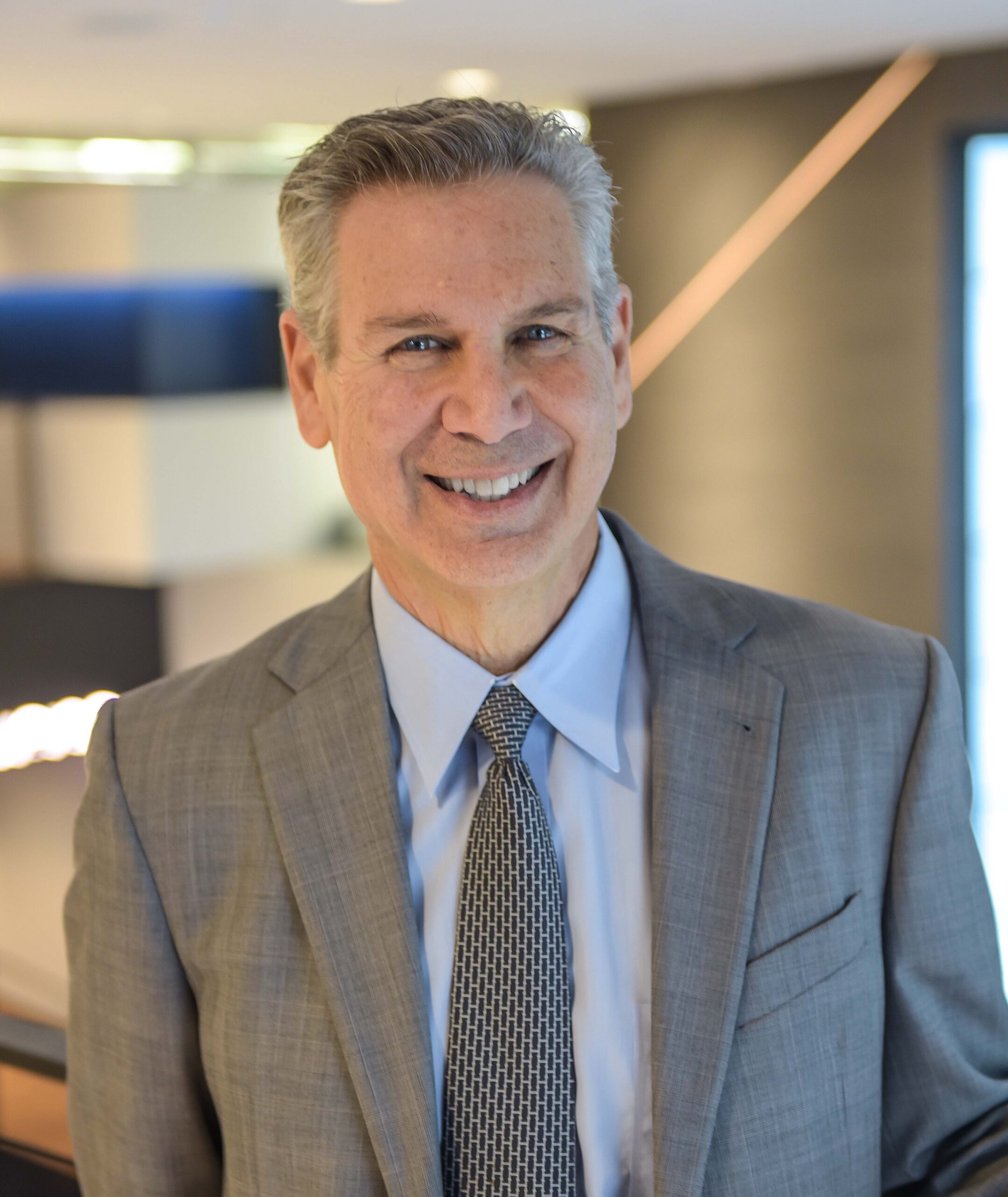 Norm Miller, a smiling man with short gray hair wears a gray suit, light blue shirt, and patterned tie, posed indoors against a softly blurred modern office background.
