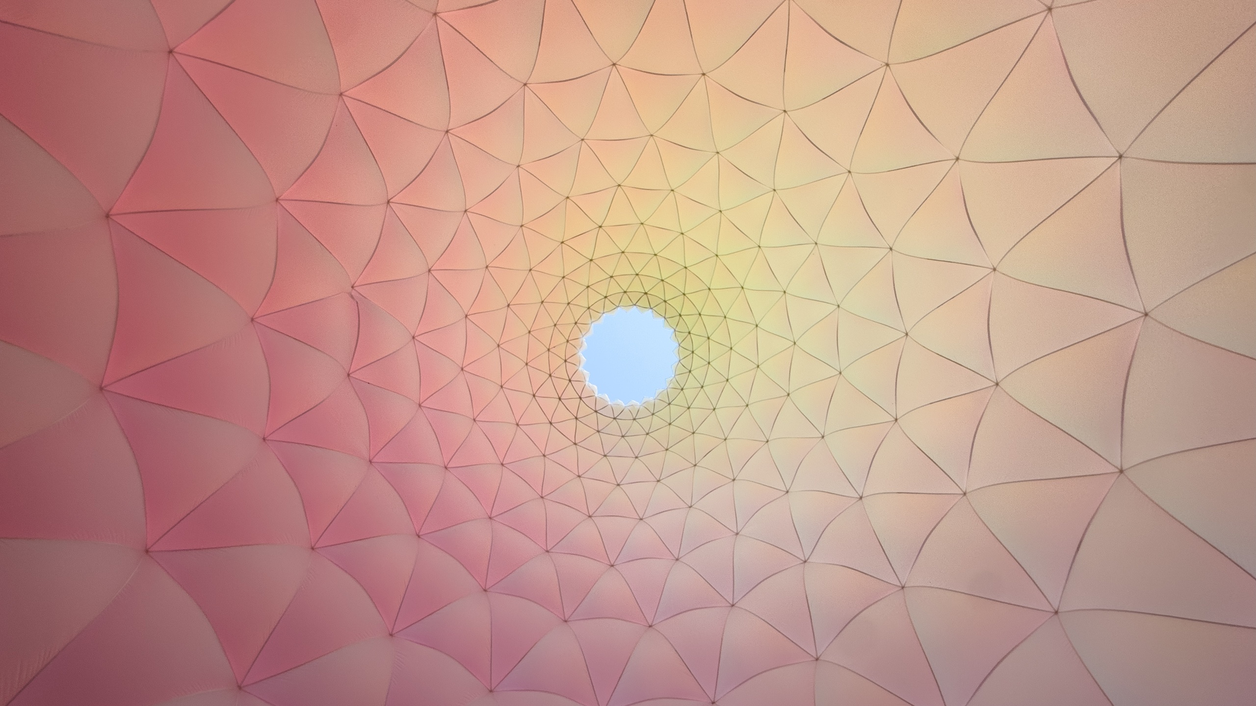 An upward view inside a translucent fabric structure, where curved geometric panels form a web-like pattern converging around a circular opening to the blue sky. Soft pink, peach, and yellow light washes across the fabric.