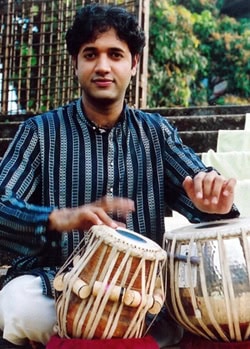 An image of Nitin Mitta seated outdoors, playing a pair of tabla drums.