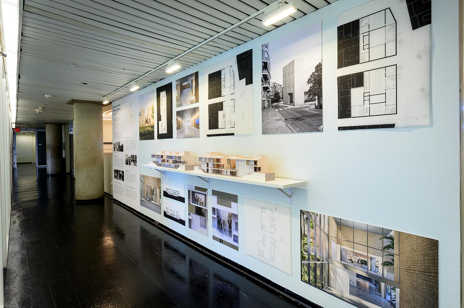 An overview of the exhibition in the hallway. The exhibition displays a series of renderings and drawings as well as two halves of a model on a long shelf showing the inside of the building.