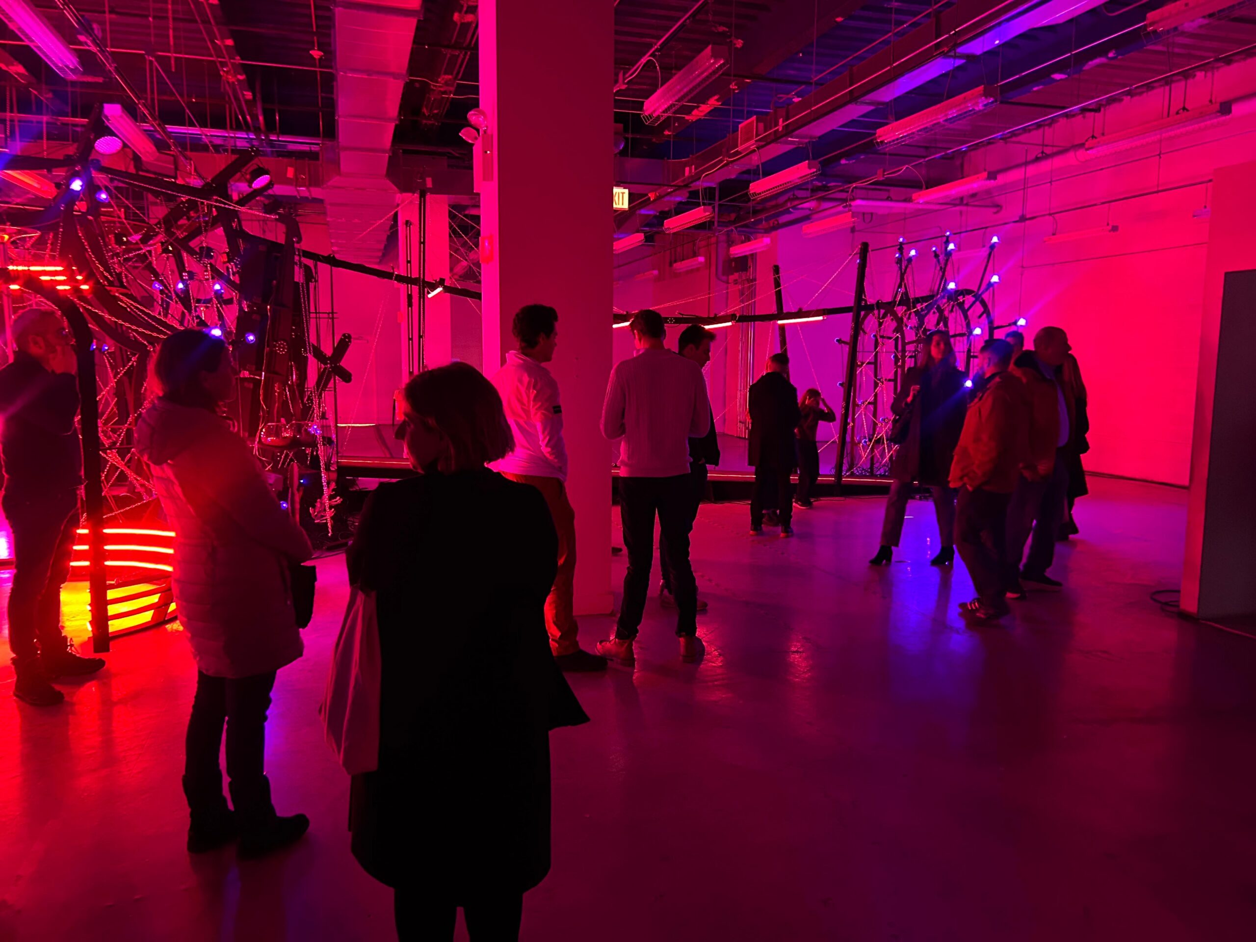 People mingling in a large industrial-style room illuminated with vivid pink, red, and blue lights around an elaborate installation.
