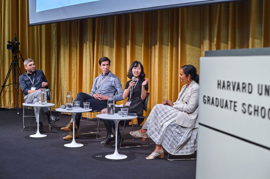 Four people site in a row as part of a panel discussion. A woman in the center holds a microphone.