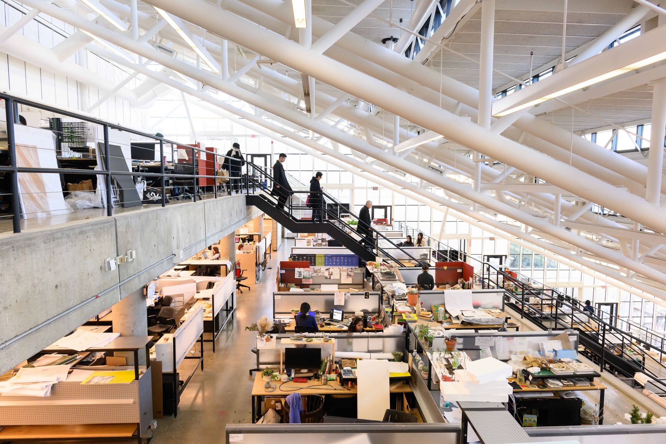 The trays in Gund Hall featuring drafting tables and computer workstations, where several people work beneath large white structural beams and beside a central staircase.