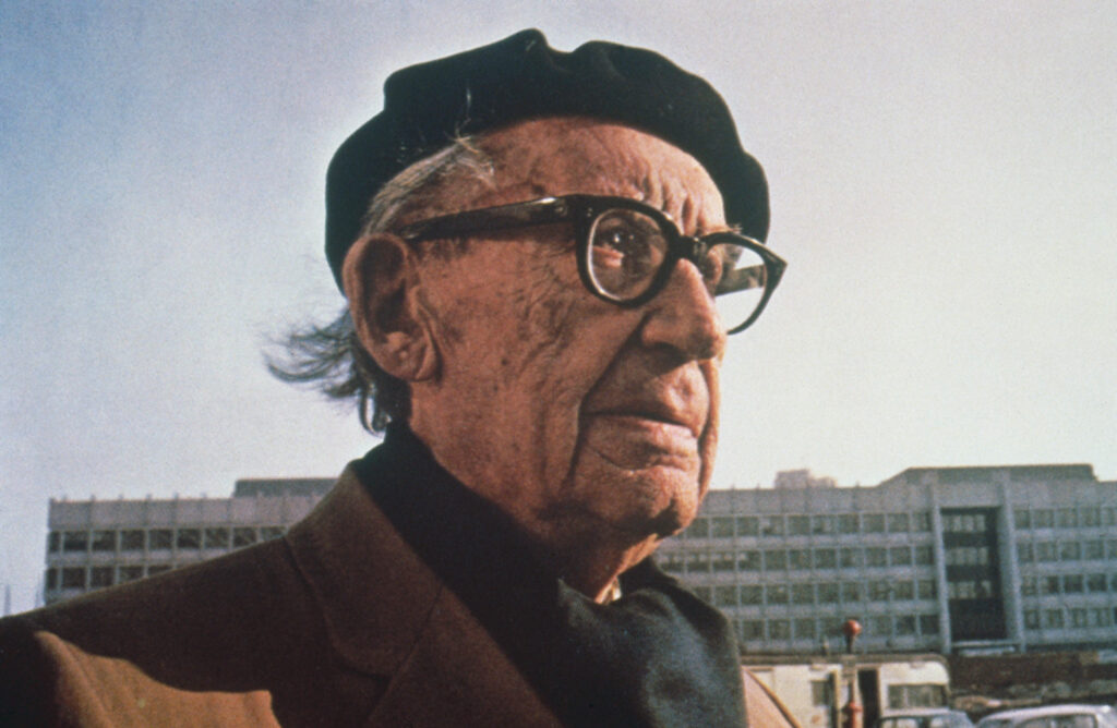 Close-up of an older man in a hat in front of a large urban building.