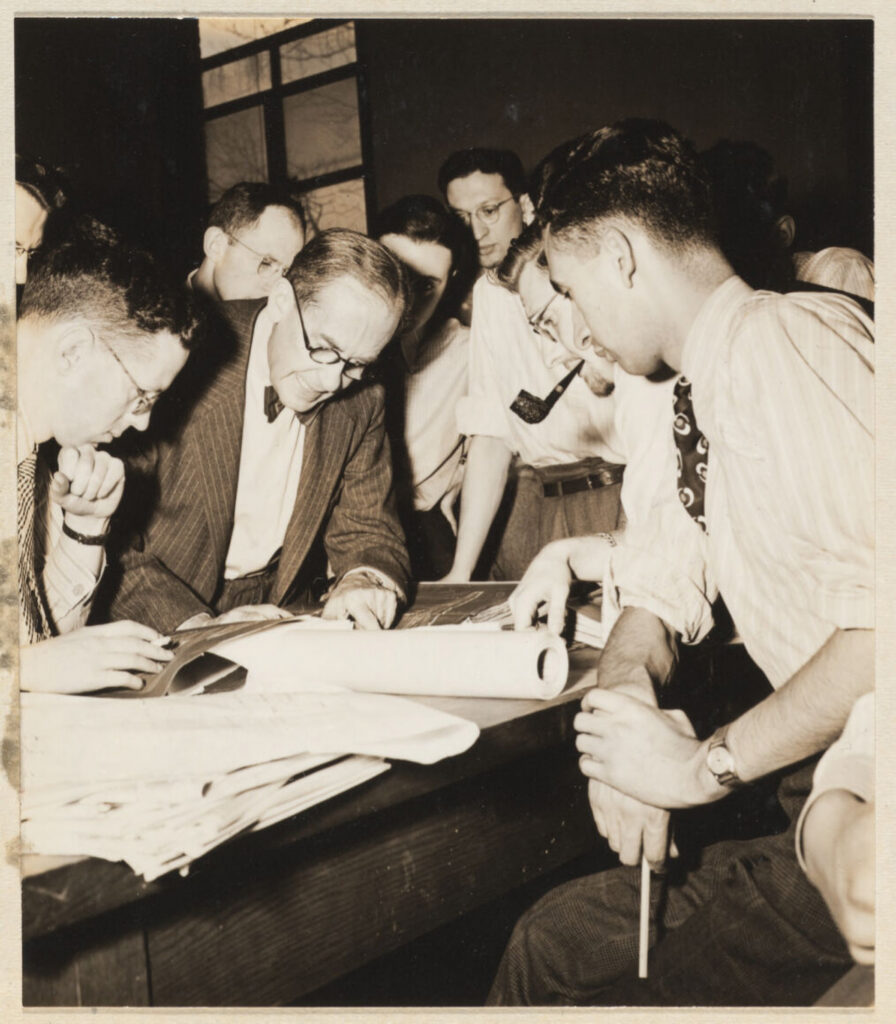 Man drawing at a desk while students gather around him.