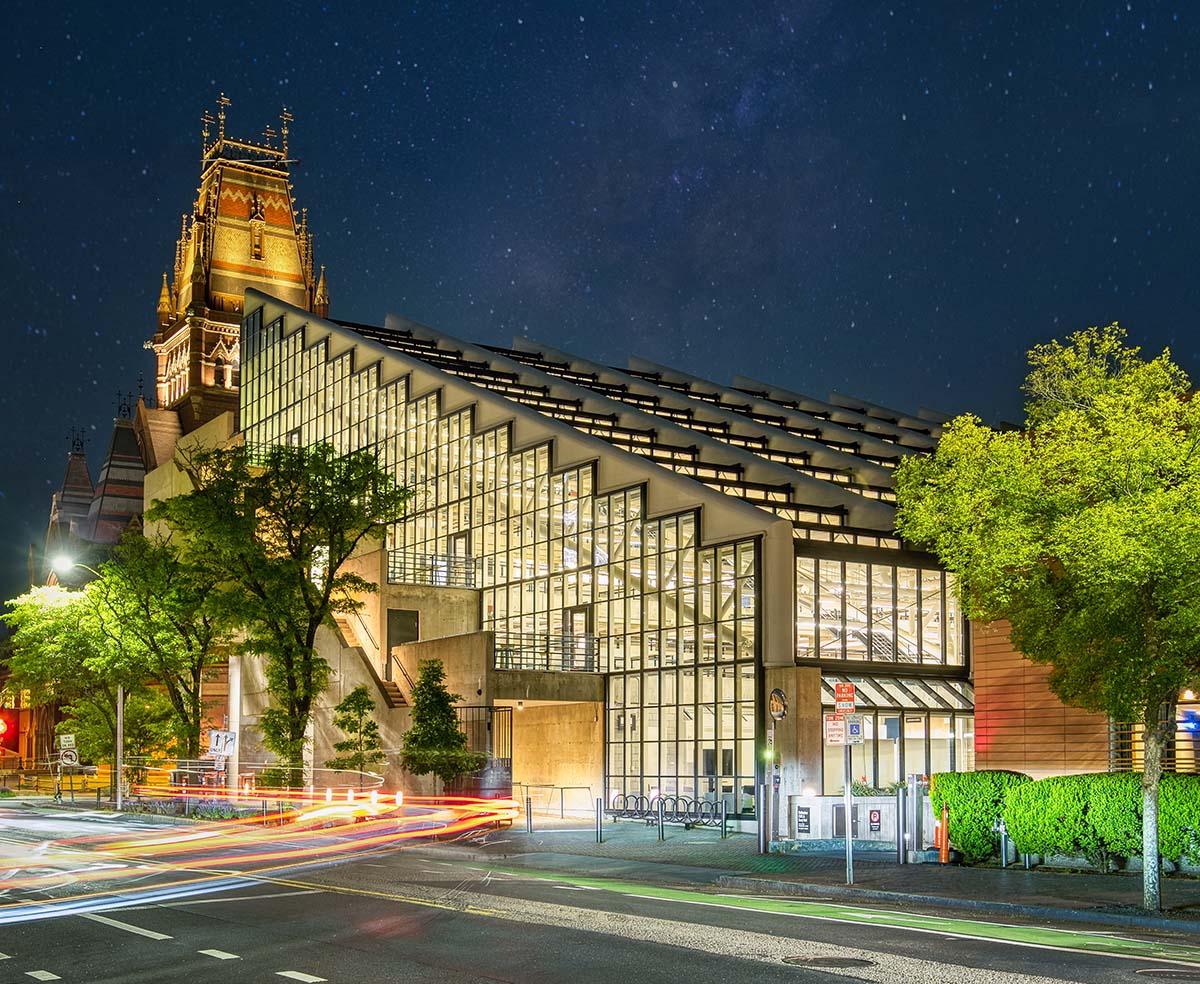 Gund Hall at night, illuminated behind trees and streaks of passing vehicle lights on the street.