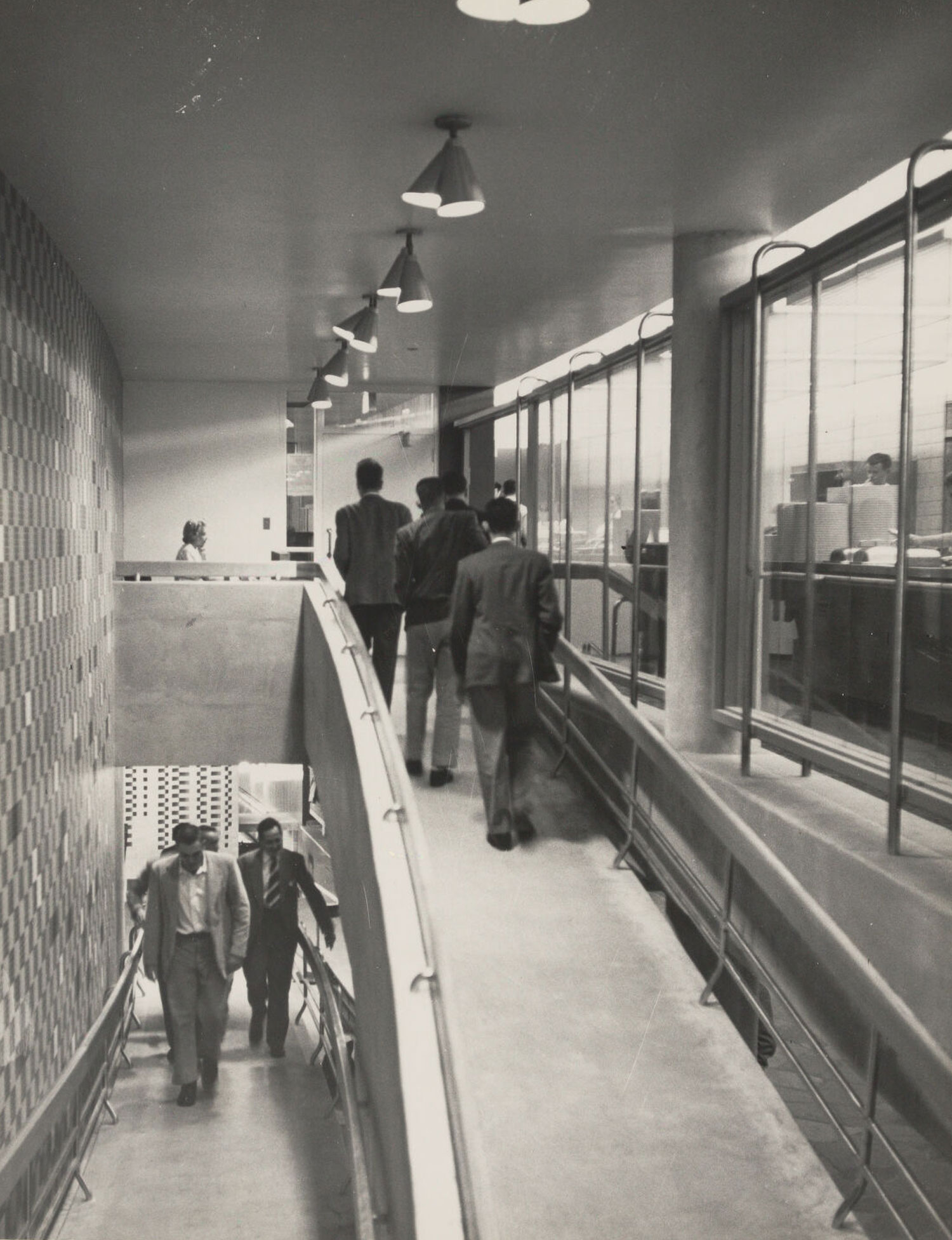 Men walking up a ramp between floors with a mural on one wall, glass on the other, and a brick sculpture at the bottom.