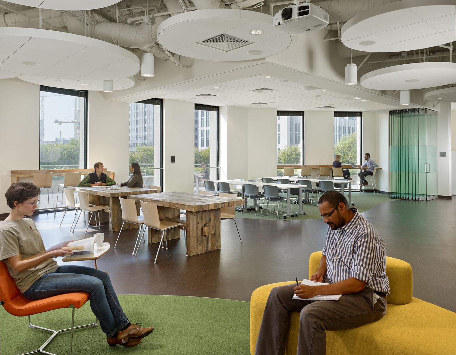 Open office with green rug, white hanging lights, wood floor, colored furniture.