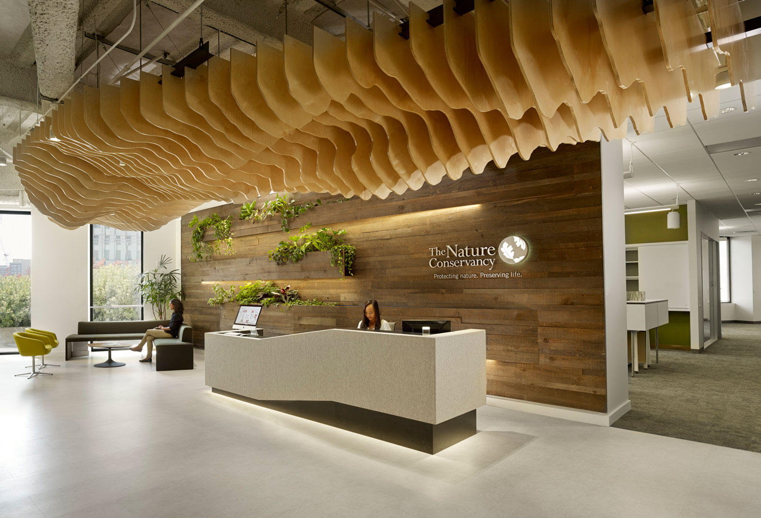 Lobby of office with textured wood ceiling and wood paneled wall behind reception desk with accents of greenery.