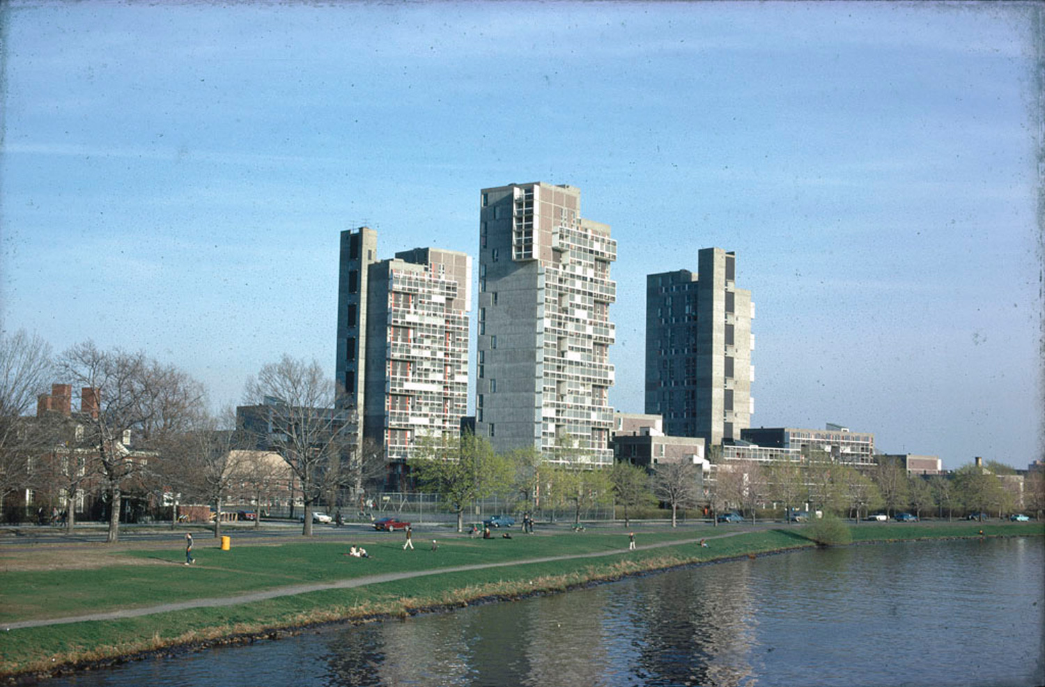 Three tall concrete buildings along a river and grassland with motorway.