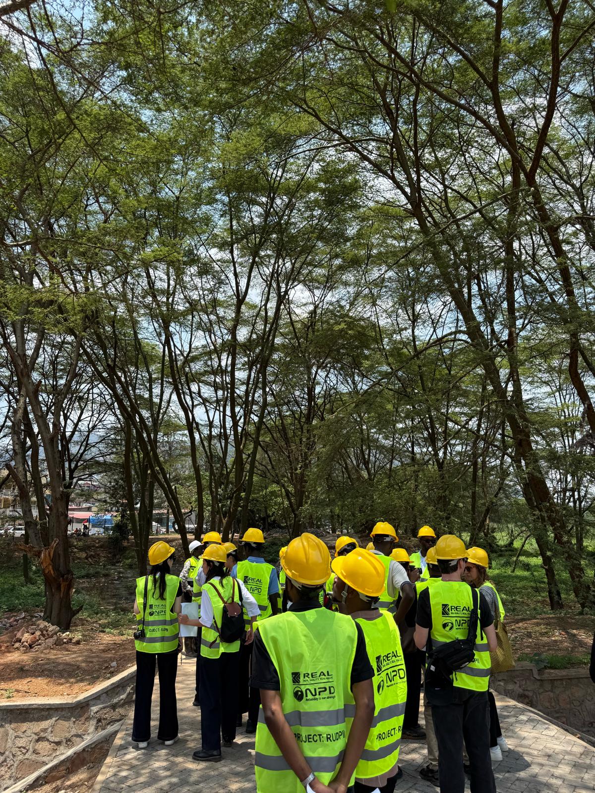 A group of people wearing yellow hard hats and neon safety vests gathers on a paved path beneath trees at an outdoor project site.