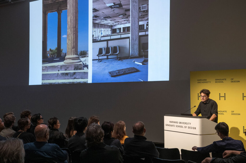 A man speaks at a podium at the GSD while a nearby screen shows two images of ruins.