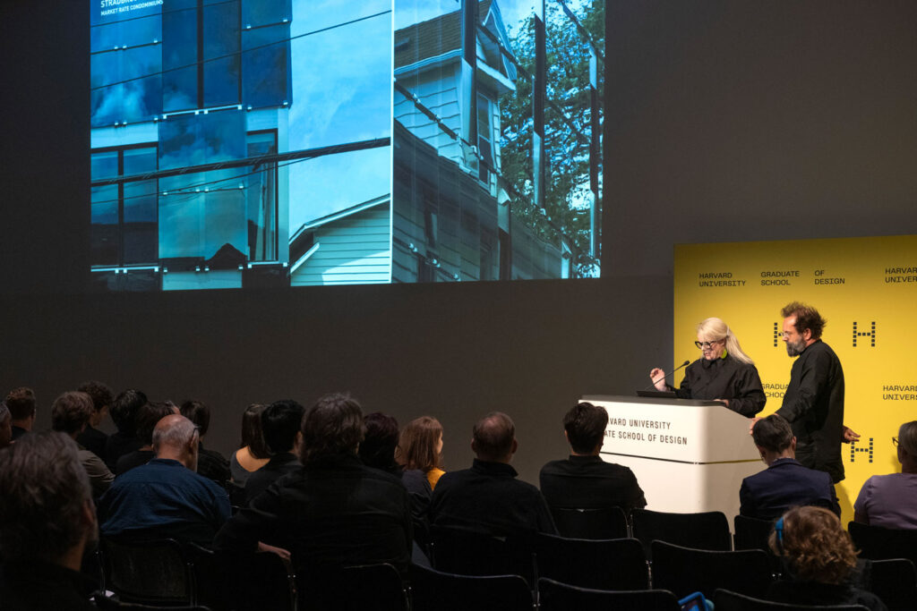 Two people speak at a podium while an image on screen displays the distorted image of a house.