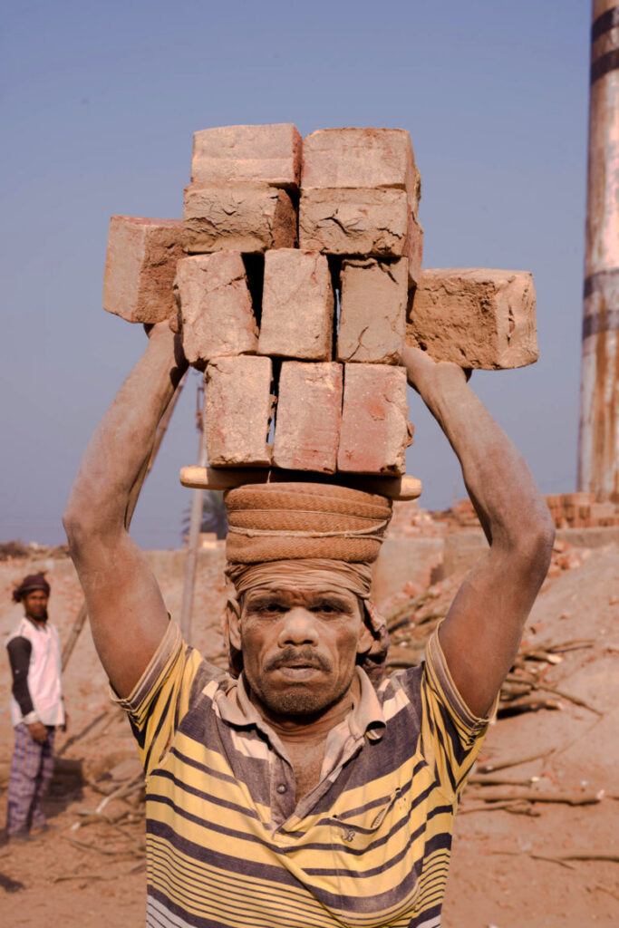A man carries several bricks on his head while looking directly at the camera. 