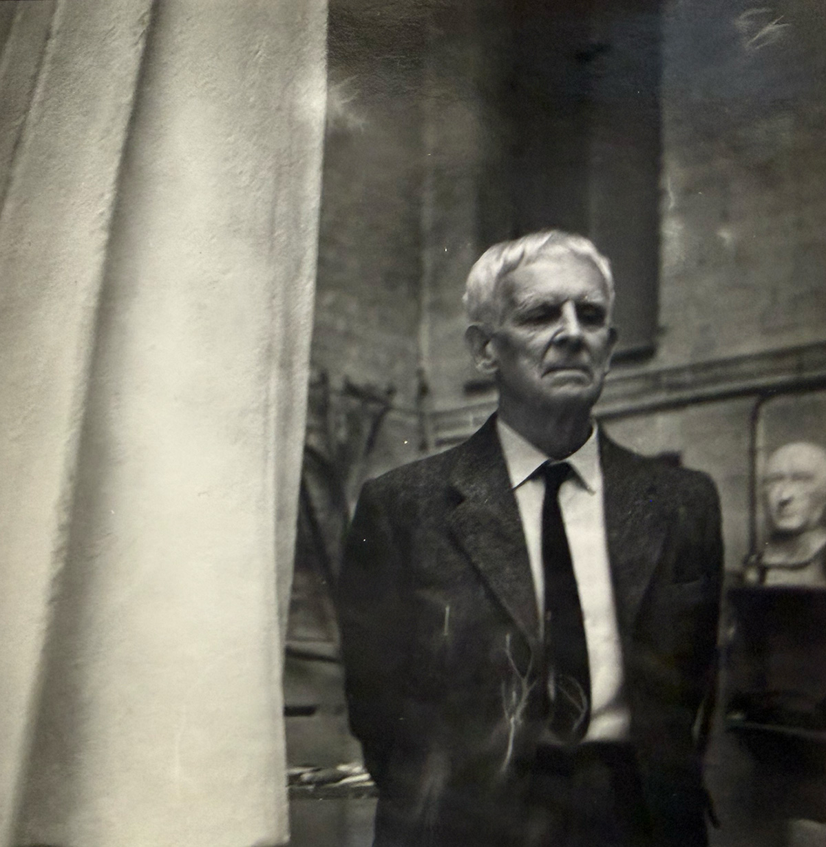 A man in a suit and tie stands next to a draped curtain, with a bust in the background.
