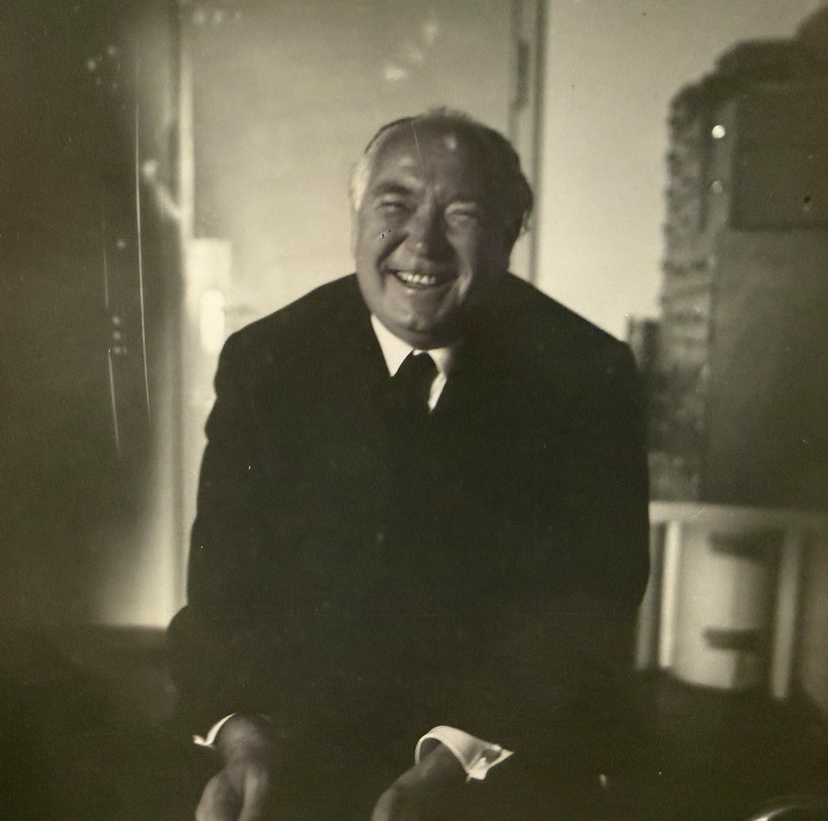 a man in a dark suit, white shirt, and dark tie, laughs, facing the camera, shoulders hunched up