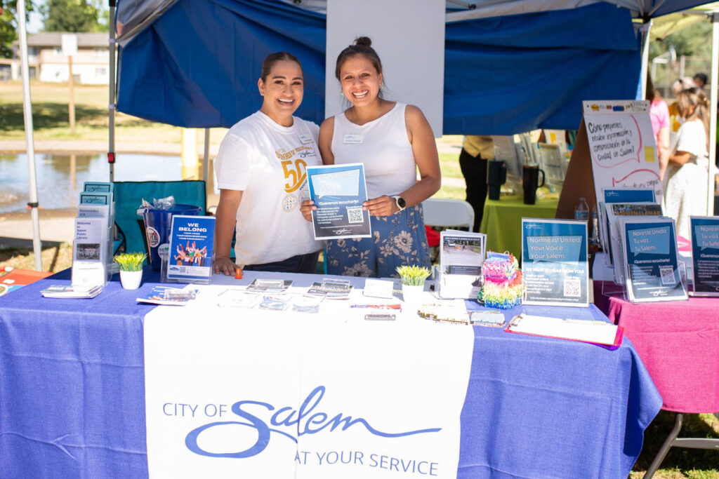 Two smiling women stand behind a City of Salem table at an outdoor event, holding up a flyer that reads “Noreste Unido. Tu voz. Tu Salem. ¡Queremos escucharte!” with a QR code. The table holds brochures, bilingual signs promoting the Northeast United survey, and small potted plants.