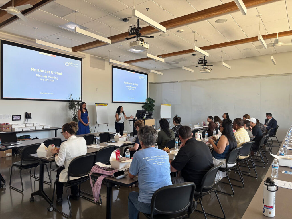 Two women present at the front of a bright meeting room to about two dozen attendees seated at long tables with boxed lunches and water bottles. Two screens display the opening slide: “Northeast United Kick-off meeting, July 29th, 2026, City’s Manager Office.”