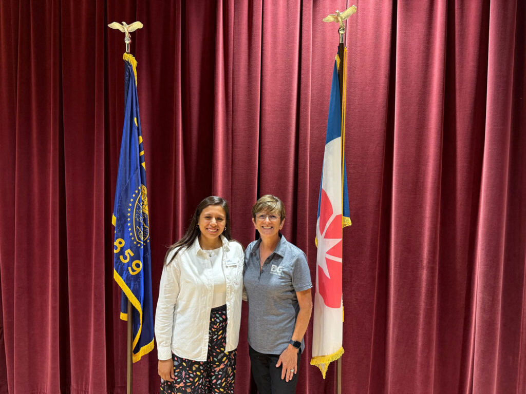 Two smiling women stand together in front of a red velvet stage curtain, between the Oregon state flag and a red, white, and blue flag, each on a pole topped with a gold eagle.
