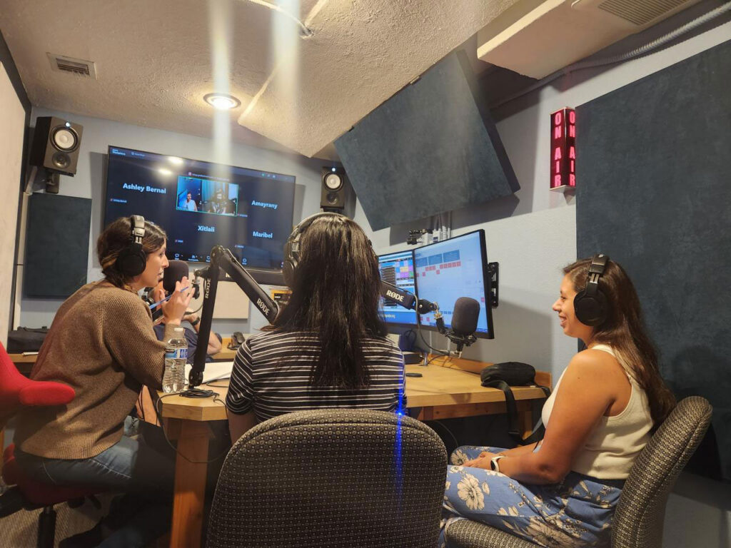 Three women wearing headphones speak into microphones in a small radio studio, with an “On Air” sign glowing red on the wall. A large screen behind them shows remote participants joining by video call.